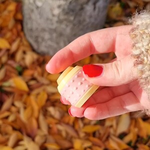 May include: A close-up of a small, pink and yellow plastic spool with a textured surface. The spool is being held by a hand with red nail polish.