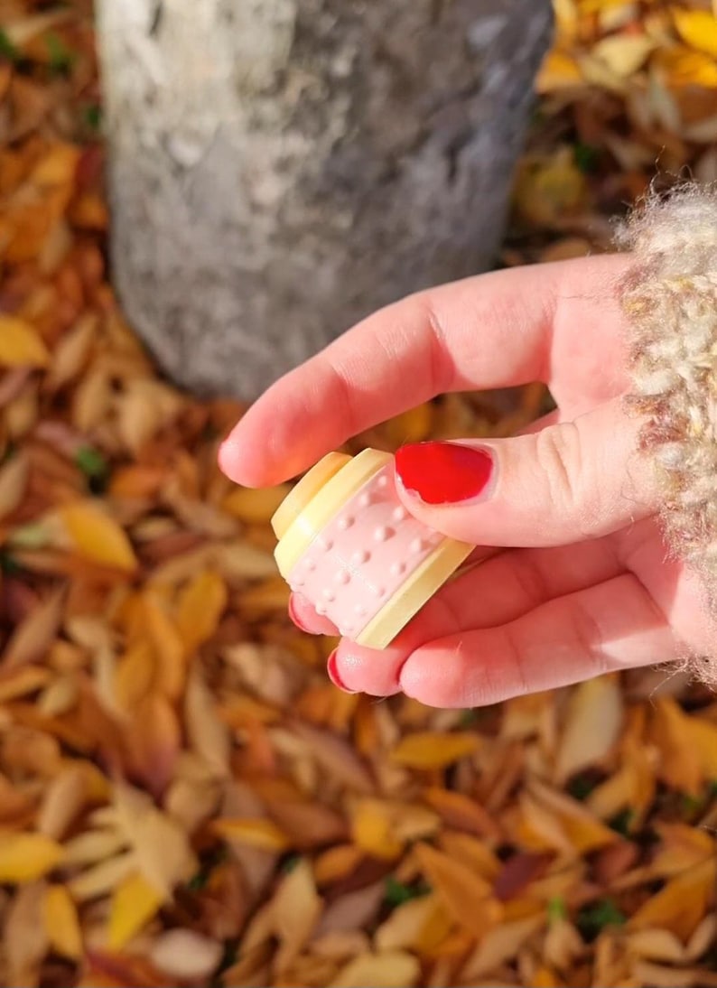May include: A close-up of a small, pink and yellow plastic spool with a textured surface. The spool is being held by a hand with red nail polish.
