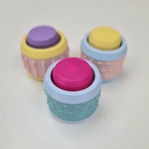 May include: Three small, colorful containers with textured lids. The containers are stacked on top of each other. The top container is teal with a pink lid, the middle container is pink with a yellow lid, and the bottom container is yellow with a purple lid.