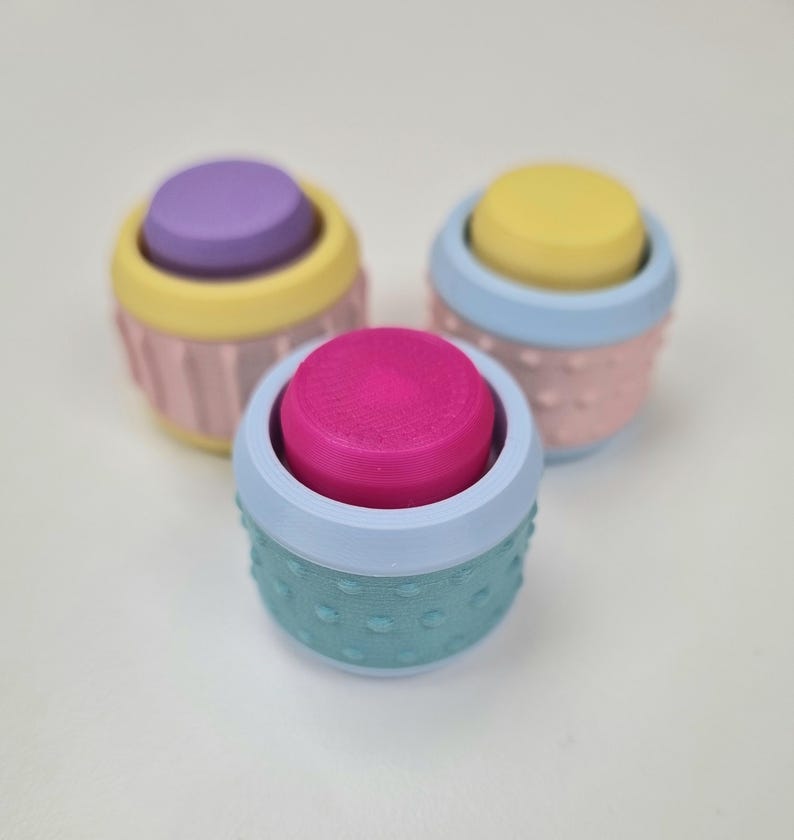 May include: Three small, colorful containers with textured lids. The containers are stacked on top of each other. The top container is teal with a pink lid, the middle container is pink with a yellow lid, and the bottom container is yellow with a purple lid.