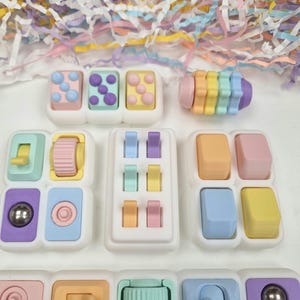 Könnte beinhalten: Eine Sammlung farbenfroher Zappelspielzeuge in verschiedenen Formen und Designs. Die Spielzeuge weisen Pastellfarben wie Rosa, Blau, Gelb und Lila auf. Einige haben Knöpfe, Rollen und Kugeln. Der Hintergrund umfasst farbiges geschreddertes Papier.