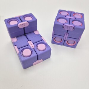 May include: Two purple and light pink fidget cubes constructed from multiple hinged square segments. Each segment features circular indentations and raised buttons, allowing the cubes to be folded and manipulated into various geometric shapes.