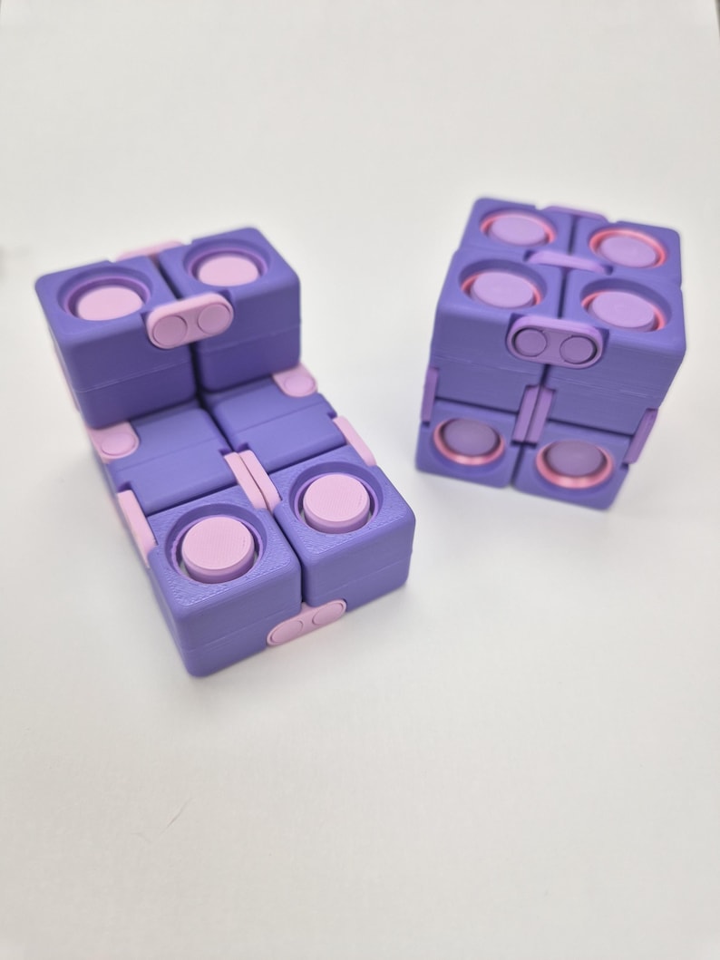 May include: Two purple and light pink fidget cubes constructed from multiple hinged square segments. Each segment features circular indentations and raised buttons, allowing the cubes to be folded and manipulated into various geometric shapes.