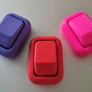 May include: Three rectangular rocker switches in purple, red, and bright pink. Each switch features a raised central button surrounded by a smooth, rounded frame. The purple switch has a subtle diagonal line pattern on its surface.