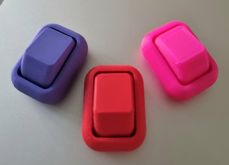 May include: Three rectangular rocker switches in purple, red, and bright pink. Each switch features a raised central button surrounded by a smooth, rounded frame. The purple switch has a subtle diagonal line pattern on its surface.