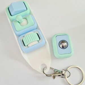 Puede incluir: Un llavero blanco con tres botones de colores pastel y un bot&oacute;n a juego separado. Los botones son de color azul claro y verde menta. El llavero tiene un cierre plateado y un llavero plateado.