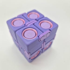 May include: A purple and light pink fidget cube toy featuring a modular design with multiple hinged sections. Each face of the cube displays a circular, textured button element surrounded by a light pink ring, constructed with a visible 3D printed finish.
