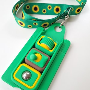 Puede incluir: Un juguete de rompecabezas verde y amarillo con un cord&oacute;n de girasol a juego. El rompecabezas tiene tres secciones con diferentes formas y colores, incluyendo una bola, un bot&oacute;n y un cilindro. El cord&oacute;n es verde con girasoles amarillos.