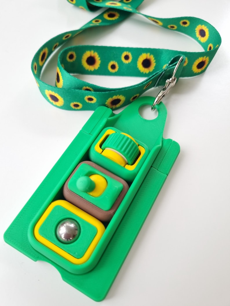 Puede incluir: Un juguete de rompecabezas verde y amarillo con un cord&oacute;n de girasol a juego. El rompecabezas tiene tres secciones con diferentes formas y colores, incluyendo una bola, un bot&oacute;n y un cilindro. El cord&oacute;n es verde con girasoles amarillos.