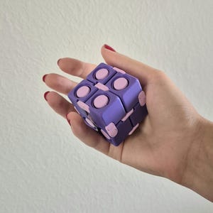 May include: A purple and light pink fidget cube toy featuring multiple interconnected square segments with circular buttons on each face. The cube is constructed from a series of hinged parts that allow it to fold and unfold into different shapes.