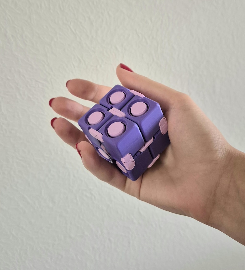 May include: A purple and light pink fidget cube toy featuring multiple interconnected square segments with circular buttons on each face. The cube is constructed from a series of hinged parts that allow it to fold and unfold into different shapes.