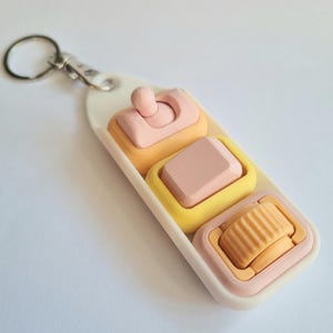 May include: A keychain featuring a white rectangular base with three interactive components: a pink toggle switch, a pink square keycap, and an orange ridged scroll wheel, all set within yellow and pink frames on a white plastic mount.