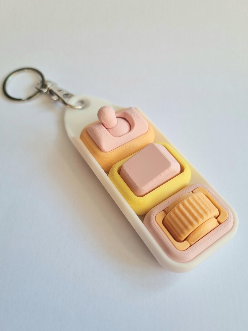 May include: A keychain featuring a white rectangular base with three interactive components: a pink toggle switch, a pink square keycap, and an orange ridged scroll wheel, all set within yellow and pink frames on a white plastic mount.