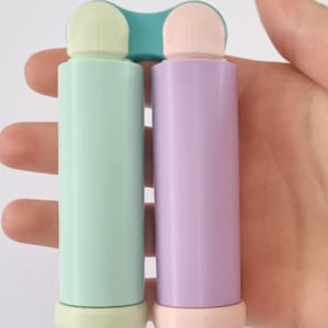 Könnte beinhalten: Ein Kunststoffbehälter mit zwei Kammern, bestehend aus einem hellen mintgrünen Zylinder auf der linken Seite und einem hellen lavendelfarbenen Zylinder auf der rechten Seite. Beide Zylinder sind oben durch eine türkise Brücke verbunden.