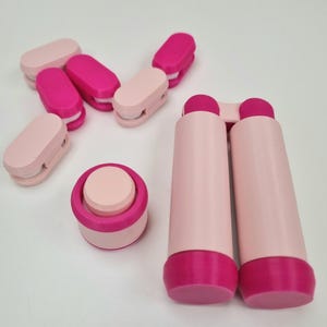 May include: A set of pink and magenta phone stand accessories. The set includes a small round stand and two long, cylindrical stands.