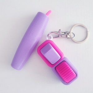 Pode incluir: Um chaveiro roxo com um interruptor de alternância rosa e roxo. O chaveiro tem um anel prateado e um clipe prateado.