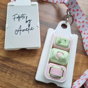Può includere: Due giocattoli fidget bianchi con dettagli rosa e verdi. Uno ha il testo "Fidgets by Amelie" in scrittura nera. Sono attaccati a un cordino floreale con un motivo rosa e bianco. Lo sfondo è una superficie di legno.