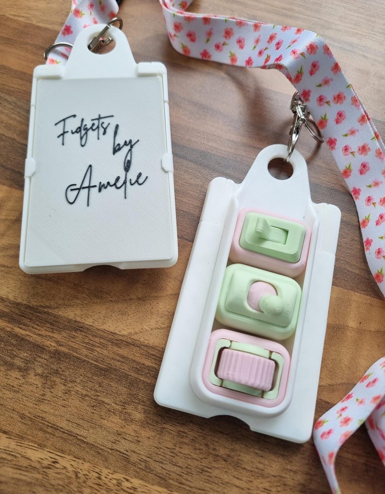 Puede incluir: Juguete blanco para inquietos con tres mecanismos de empujar y hacer clic en colores pastel. El juguete est&aacute; sujeto a un cord&oacute;n con estampado floral. El texto "Fidgets by Amelie" est&aacute; impreso en un juguete blanco separado.