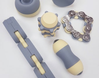 May include: A collection of fidget toys in various shapes and designs. The toys are primarily gray and cream-colored, including a chain, a segmented link, and several rounded and geometric shapes. The objects are arranged on a white surface.