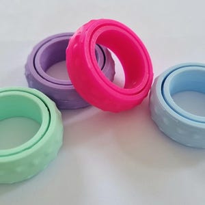 May include: Four colorful fidget rings in shades of green, purple, pink, and blue. Each ring has a textured outer band and a smooth inner ring. The rings are designed to spin and provide tactile stimulation.