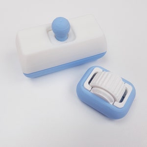 May include: Two small, blue and white plastic objects. One is a rectangular block with a white top and a blue bottom. The top has a small white knob. The other object is a small, blue, rectangular block with a white, ridged wheel on top.