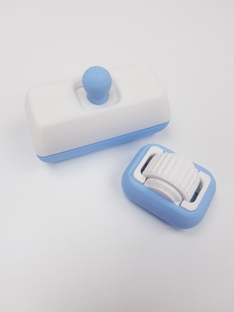 May include: Two small, blue and white plastic objects. One is a rectangular block with a white top and a blue bottom. The top has a small white knob. The other object is a small, blue, rectangular block with a white, ridged wheel on top.