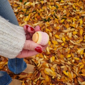 May include: A pink and yellow patterned lip balm tube with a white cap. The lip balm is being held in a hand with red nail polish.