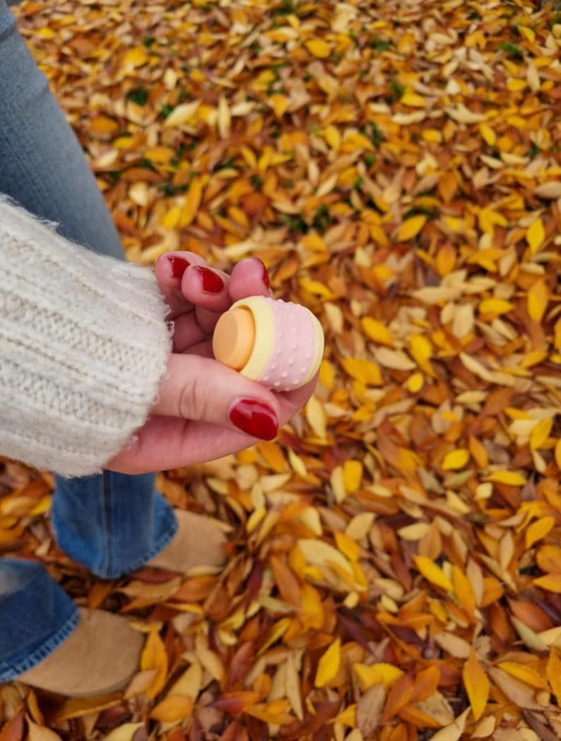May include: A pink and yellow patterned lip balm tube with a white cap. The lip balm is being held in a hand with red nail polish.