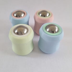 May include: Four pastel-coloured rollerball applicators. Each applicator has a rounded base in shades of light blue, pink, green, and yellow. A silver metal ball sits at the top of each applicator, surrounded by a contrasting ring.