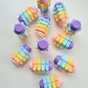 Puede incluir: Una colección de engranajes de juguete apilables de colores pastel. Los engranajes están hechos de un material suave y flexible y están diseñados para apilarse uno encima del otro. Los colores de los engranajes son morado, azul, verde, amarillo y naranja.