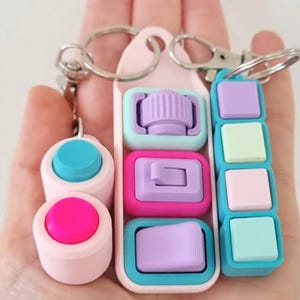 Puede incluir: Una colección de juguetes fidget de colores en llaveros. Los juguetes presentan varias formas y colores, incluyendo rosa, azul, morado y turquesa. Los llaveros son plateados y el fondo es blanco.