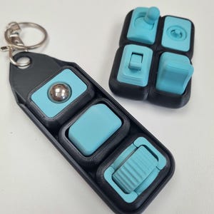Puede incluir: Un juguete fidget negro y turquesa con un llavero. El juguete presenta varios elementos t&aacute;ctiles, incluyendo una bola rodante, un bot&oacute;n y un interruptor. El dise&ntilde;o est&aacute; impreso en 3D, con una combinaci&oacute;n de colores negro y turquesa.
