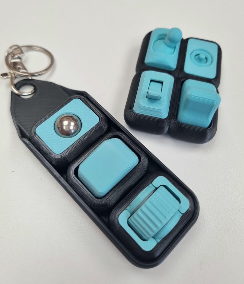 Puede incluir: Un juguete fidget negro y turquesa con un llavero. El juguete presenta varios elementos t&aacute;ctiles, incluyendo una bola rodante, un bot&oacute;n y un interruptor. El dise&ntilde;o est&aacute; impreso en 3D, con una combinaci&oacute;n de colores negro y turquesa.