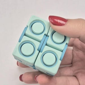 May include: A square-shaped fidget toy constructed from four smaller interlocking blocks. The toy features a light mint green exterior with circular indentations and bright blue hinges connecting the individual segments together.
