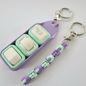 Puede incluir: Dos llaveros con una combinaci&oacute;n de colores morado y verde menta. Un llavero presenta tres juguetes fidget de color verde menta y blanco en un soporte morado. El otro llavero tiene un dise&ntilde;o de cuentas con segmentos morados y verde menta alternados.