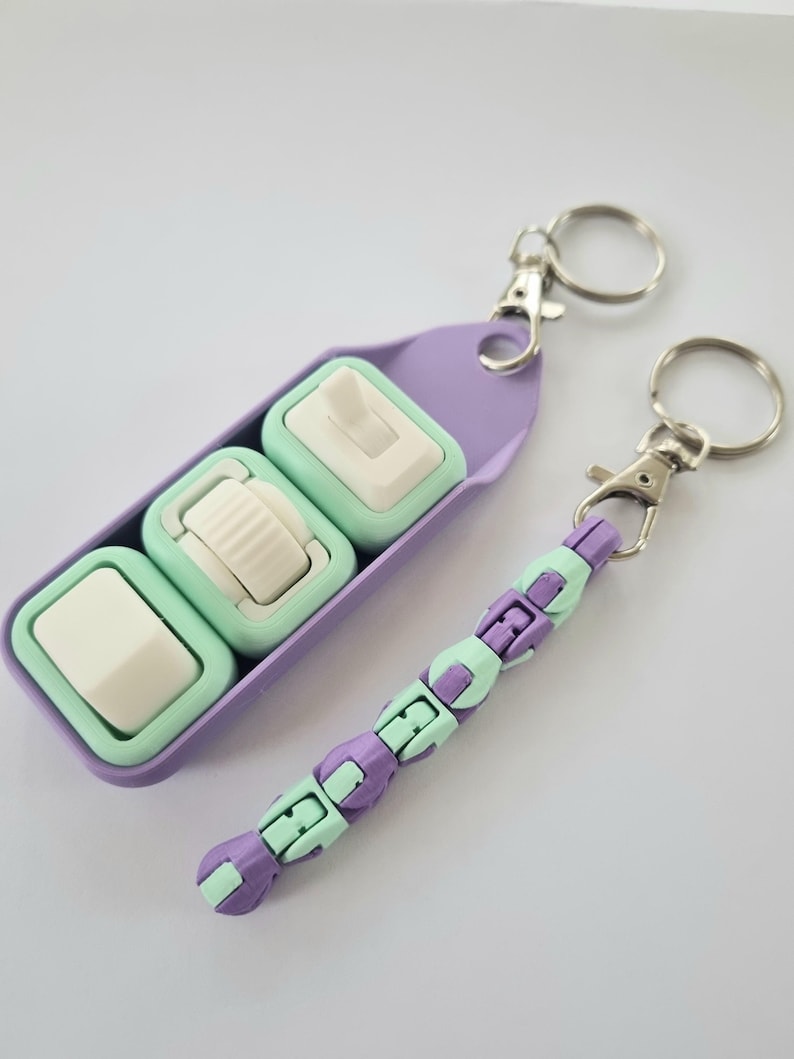 Puede incluir: Dos llaveros con una combinaci&oacute;n de colores morado y verde menta. Un llavero presenta tres juguetes fidget de color verde menta y blanco en un soporte morado. El otro llavero tiene un dise&ntilde;o de cuentas con segmentos morados y verde menta alternados.