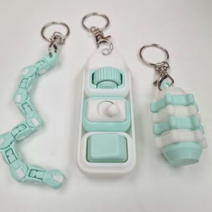 May include: Three mint green and white fidget keychains. One is a long, articulated snake-like chain, one is a rectangular controller with a roller, switch, and button, and one is a cylindrical, segmented, stackable piece, all with metal rings.