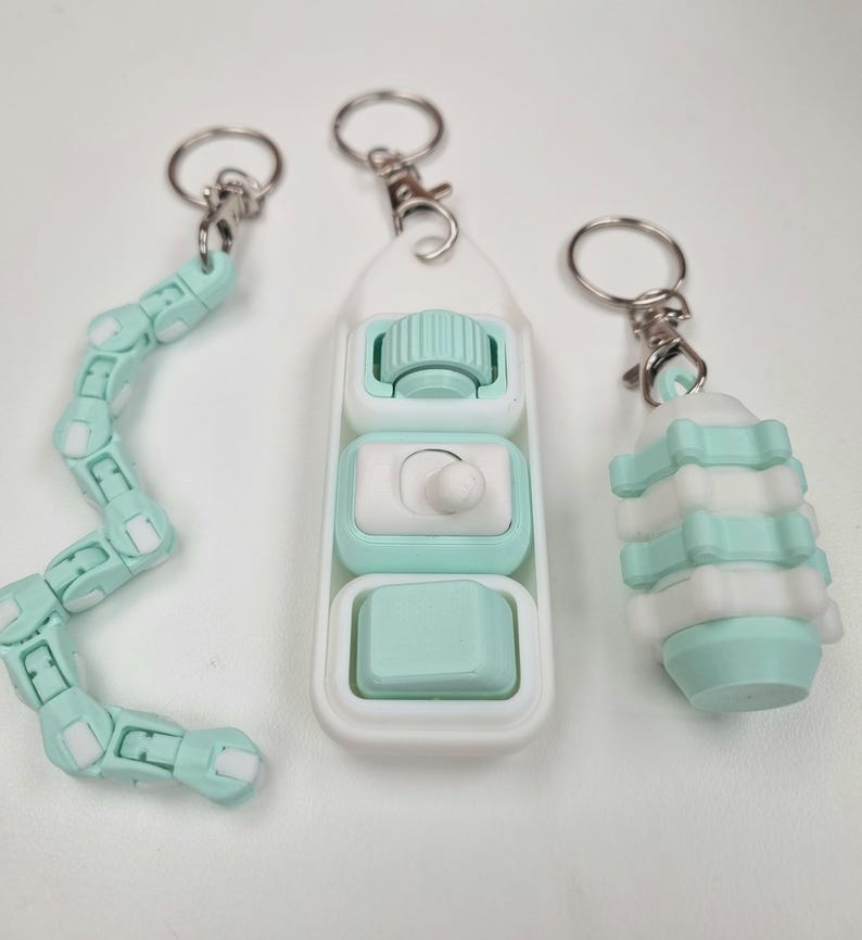 May include: Three mint green and white fidget keychains. One is a long, articulated snake-like chain, one is a rectangular controller with a roller, switch, and button, and one is a cylindrical, segmented, stackable piece, all with metal rings.