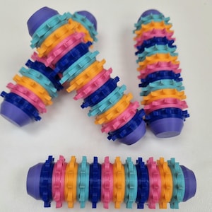 Puede incluir: Una colección de juguetes cilíndricos coloridos. Cada juguete está compuesto por engranajes apilados multicolores en tonos de azul, rosa, naranja y turquesa, con tapas moradas. Los juguetes están dispuestos sobre una superficie blanca.