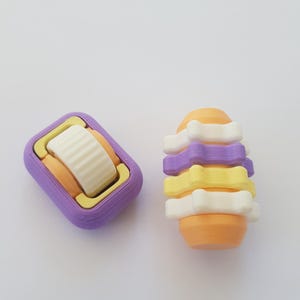May include: Two 3D printed toys, one is a purple and orange rectangle with a white wheel, the other is a stack of orange, yellow, and white rings.