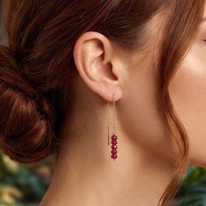 May include: A gold-toned threader earring featuring a delicate chain that passes through the earlobe, ending in a vertical row of four small, faceted, deep red circular beads that hang freely below the ear.