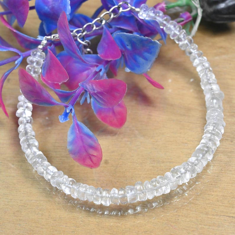 May include: A clear, faceted gemstone necklace with a silver chain clasp. The necklace is composed of small, evenly sized beads. The background features artificial purple and blue flowers, creating a contrast with the necklace's clarity.