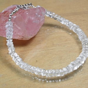 May include: A clear quartz bracelet with a silver clasp is displayed on a wooden surface. The bracelet is composed of small, rounded quartz beads. A large, rough-cut pink stone is in the background.