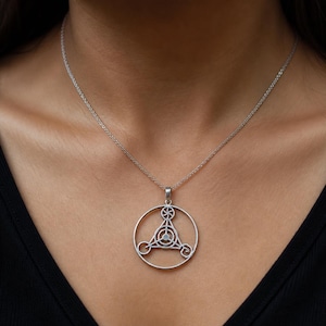 Puede incluir: Collar con colgante plateado con un diseño geométrico. El colgante presenta un triángulo dentro de un círculo, con patrones detallados y una pequeña piedra clara. El collar se lleva sobre un fondo oscuro.