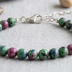 Pode incluir: Uma pulseira de contas composta por pedras de zoisite redondas e polidas com inclusões de verde vibrante e magenta profundo. As contas estão enfiadas e presas com um fecho lagosta de metal prateado e uma corrente extensora ajustável.