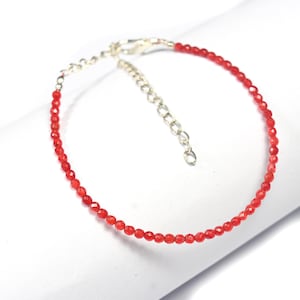 May include: A delicate red beaded bracelet with a silver-colored clasp and adjustable chain. The bracelet features small, faceted red beads strung together, creating a subtle sparkle. The bracelet is displayed on a white surface.