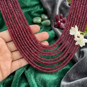 Puede incluir: Un collar de múltiples hilos con pequeñas cuentas de rubí facetadas de color rojo intenso dispuestas en cascada. El collar está adornado con una pequeña flor blanca y se exhibe junto a piedras verdes lisas y un grupo de cristales de color rojo oscuro.