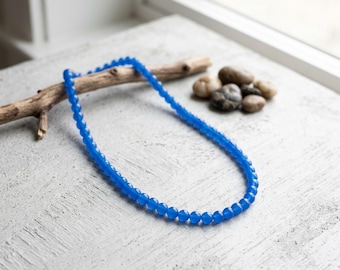 May include: A single strand necklace composed of small, uniform, round, translucent blue beads. The necklace is arranged in an oval shape, resting on a textured surface alongside a piece of driftwood and several small, smooth, grey and brown stones.