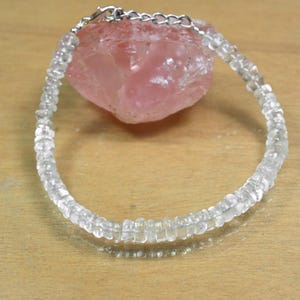 May include: A clear quartz bead bracelet with a silver-colored clasp. The bracelet is displayed on a light pink, rough-textured stone. The beads are small and round, strung together to form a delicate piece of jewelry.