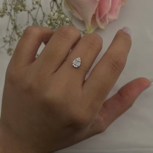 May include: A clear ring with a teardrop-shaped, faceted, clear stone set in the center. The stone is surrounded by smaller, clear stones.